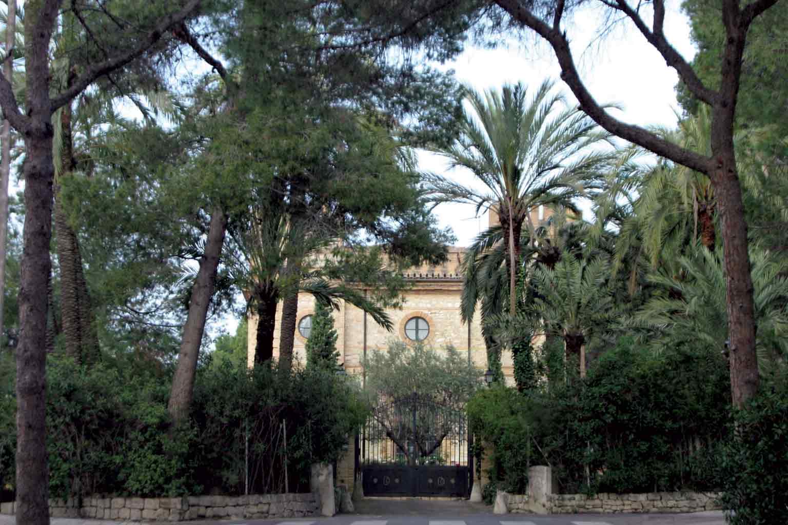 The historic baronial residence of Campolivar, a stone manor house with round windows seen through palms and pines behind a wrought-iron gate.